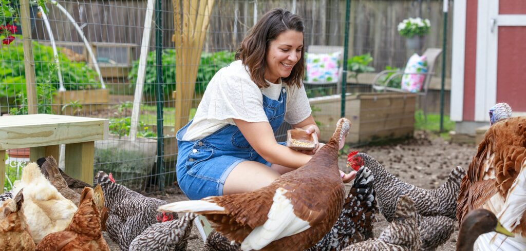 Sara Brown of Brown Family Goods on YouTube kneels in a crowd of chickens and turkeys with her hand out to feed the flock.
