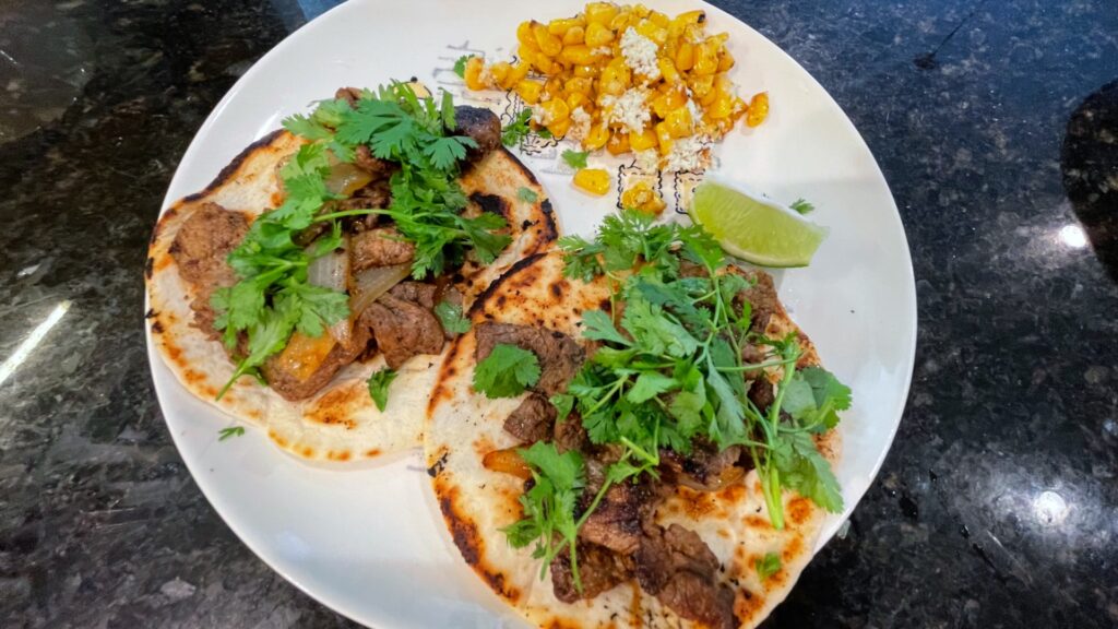 Two grilled carne asada tacos on crisped corn tortillas are served alongside wedges of lime and blackened corn cut off the cob.