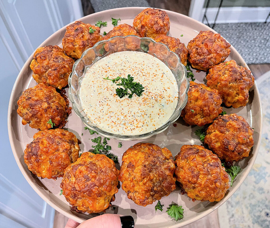 Crispy, golden brown Creole sausage balls arranged on a plate with a bowl of creamy dipping sauce in the center. Fresh parsley adds a touch of color.