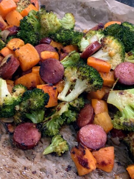 A sheet pan lined with parchment paper and filled with roasted sausage, sweet potato cubes, and broccoli florets.