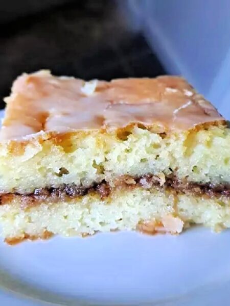A close-up of a slice of Honey Bun Cake on a white plate. The cake is layered with a brown filling and topped with a white glaze.