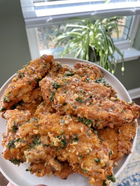 The image shows a plate of crispy, golden-brown chicken tenders. The tenders are coated in a creamy, savory sauce that appears to be garlic butter. The sauce is drizzled over the tenders and there are also some herbs sprinkled on top. The plate is resting on a table next to a window.