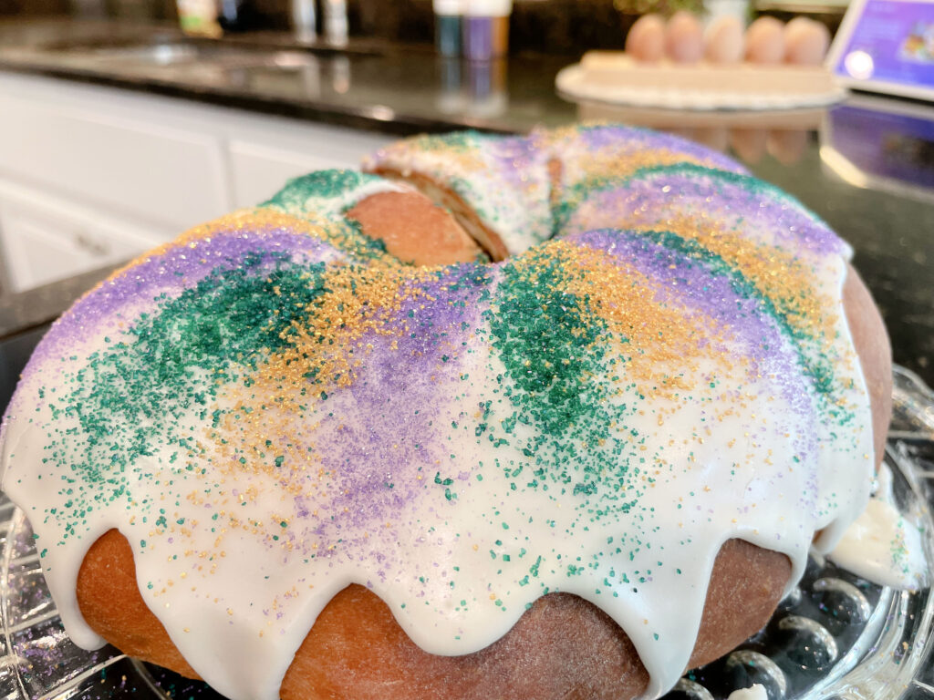 A round loaf of king cake that has been draped in white frosting has glittery purple, green and gold sparkling sugar on top.