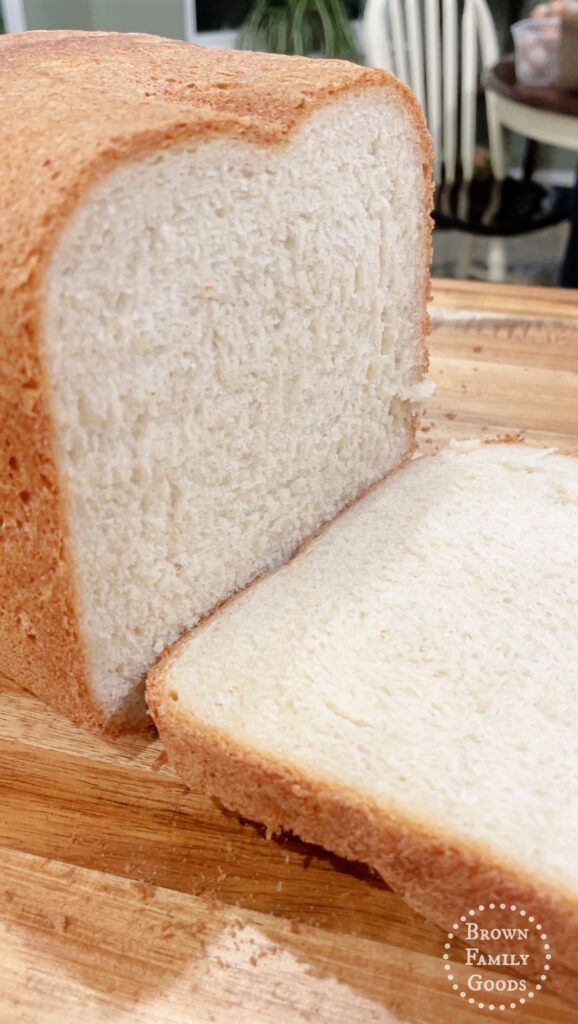 A close-up, angled view of a bread machine sourdough loaf, sliced, showing a light brown crust and a soft, white, airy interior. The bread is on a wooden cutting board. A logo for "Brown Family Goods" is visible in the bottom right corner.