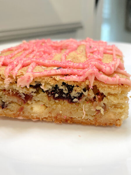 A buttery, soft baked blondie bar studded with red jammy strawberries sits on a white plate.