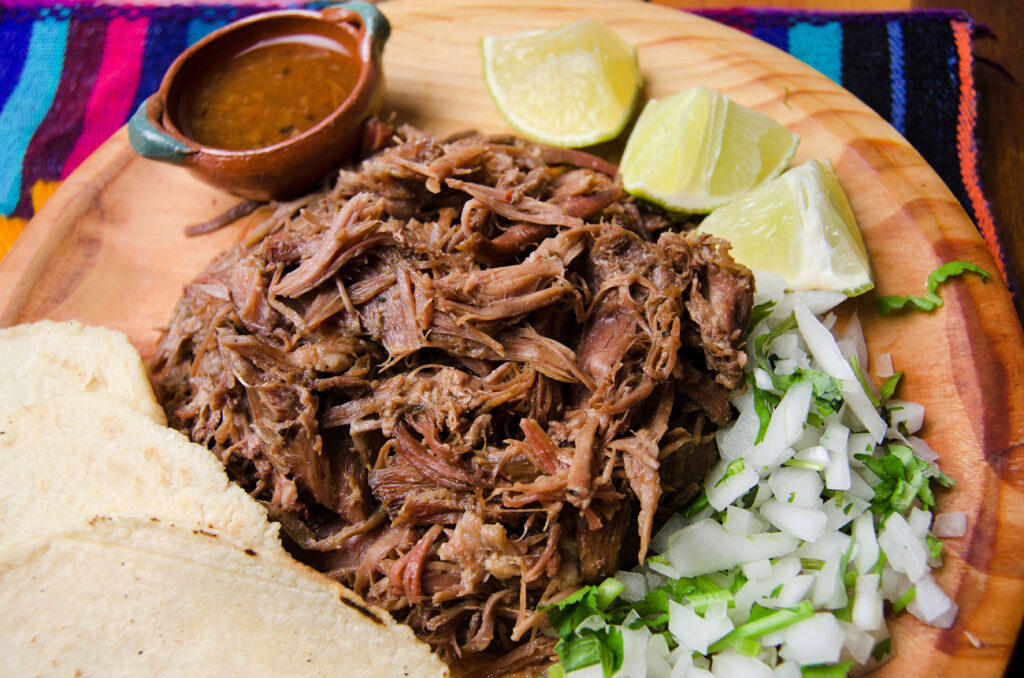 Shredded beef on a wooden plate with tortillas, lime wedges, chopped onions, and a small bowl of sauce.