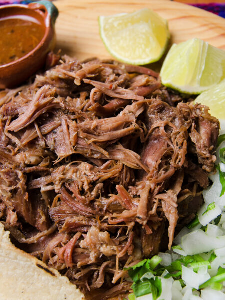 Shredded beef on a wooden plate with tortillas, lime wedges, chopped onions, and a small bowl of sauce.