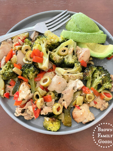 A single serving of Lemon Herb Chicken and Roasted Veggies on a gray plate with sliced avocado and pitted green olives on the side.