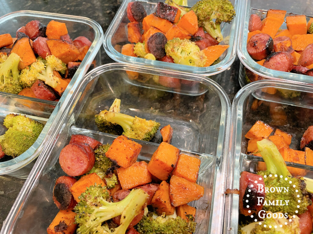 Four individual meal prep containers with two compartments, filled with servings of the Sheet Pan Sausage, Sweet Potatoes, and Broccoli.