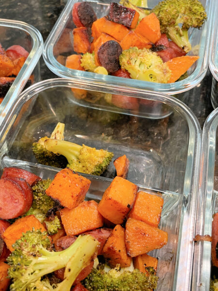 Four individual meal prep containers with two compartments, filled with servings of the Sheet Pan Sausage, Sweet Potatoes, and Broccoli.