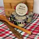 Jars of homemade Wood Butter conditioner on a red and green plaid holiday tablecloth. The jars are decorated with Christmas fabric and twine, sitting in front of a wooden cutting board and two wooden spoons.